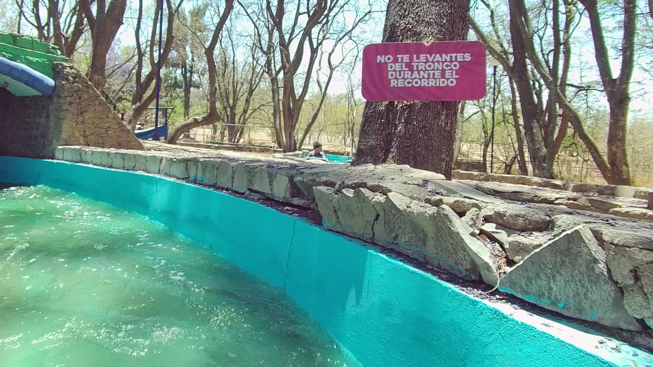 Turquoise water flows through a concrete flume in Guadalajara Mexico, with rough stone edging, tall trees, bright sky, a red warning sign, and no visible people in this frame.