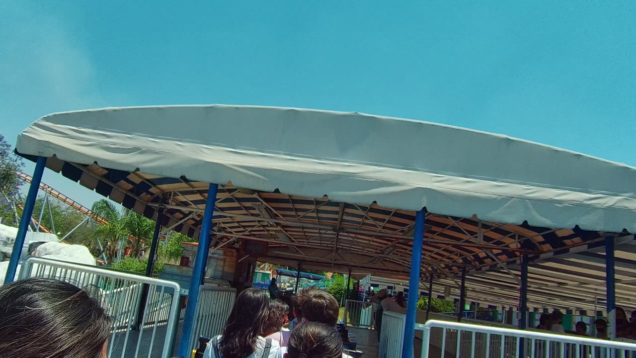 Foreground passengers sit beneath a blue and white striped canopy at a Guadalajara, Mexico transit station, with railings and shade structure visible ahead.