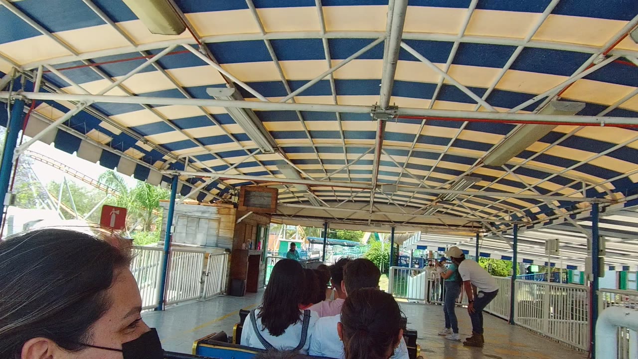 Seated passengers face a railed station area under a striped roof.