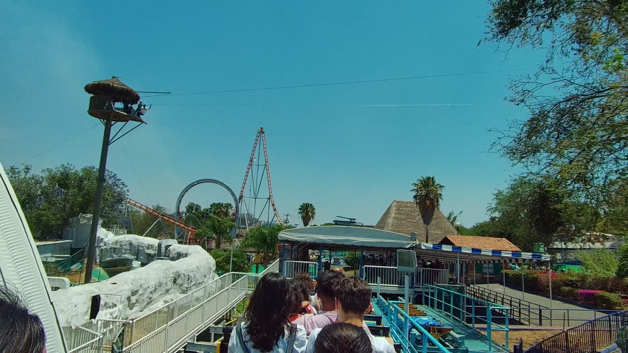 In Guadalajara Mexico, riders move past white rockwork and blue railings with a covered station, palm tree, trees, and looping roller coaster elements visible in the background.