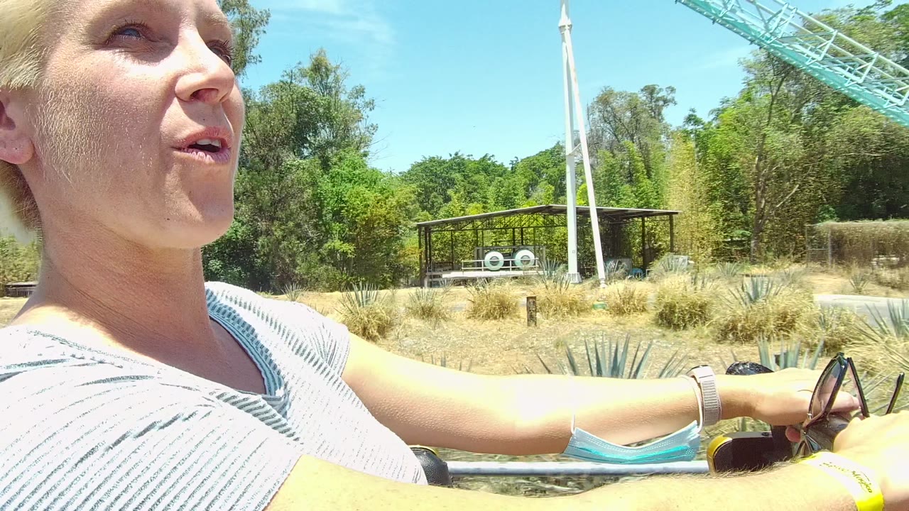In Guadalajara Mexico, a blond woman in a striped shirt rides in a vehicle with trees, dry grasses, a crane, and a blue sky behind her.