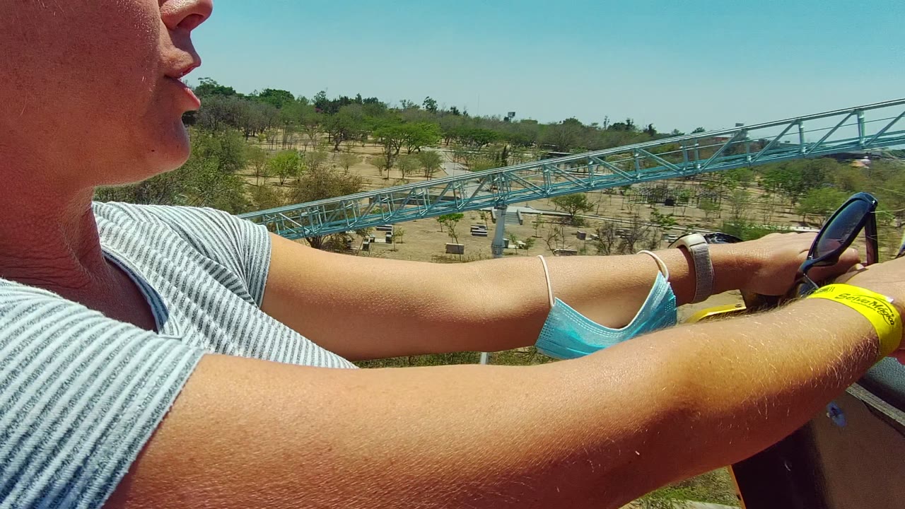 In Guadalajara Mexico, a close seat-level view shows a rider's striped shirt, arms, yellow wristband, black safety bar, blue roller coaster track, trees, and clear sky.