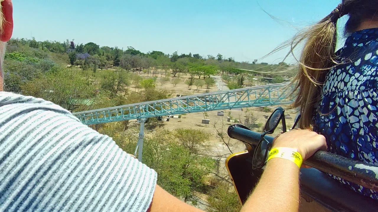 Riders beside a blue roller coaster track over trees and dry park ground