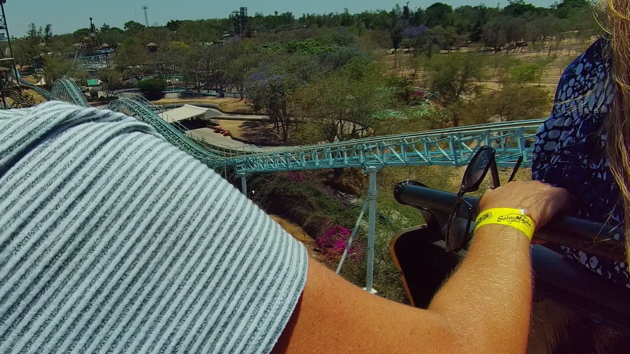 In Guadalajara Mexico, a rider in a striped shirt fills the foreground while another passenger sits ahead, with blue track, ride supports, trees, and park structures visible below.