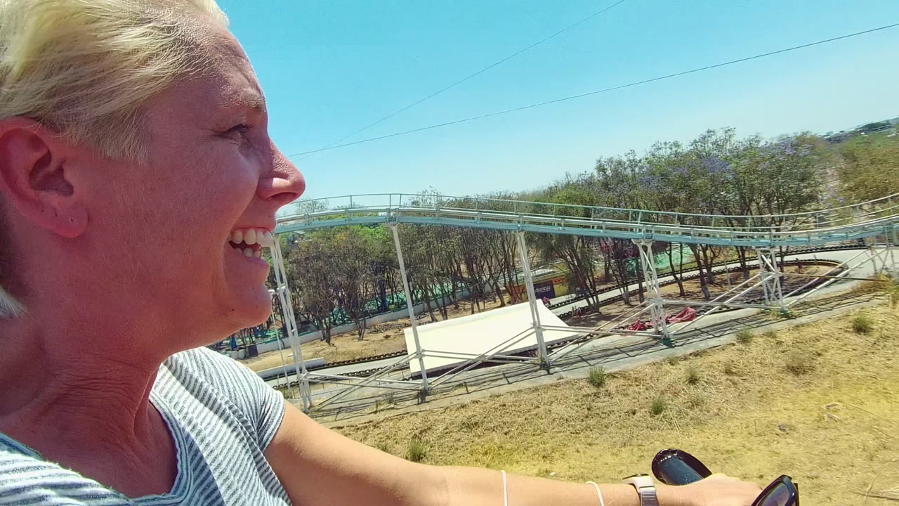 At 0:00, a woman smiles in profile on a roller coaster in Mexico, with pale blue track, dry ground, scattered trees, and clear sky behind her.
