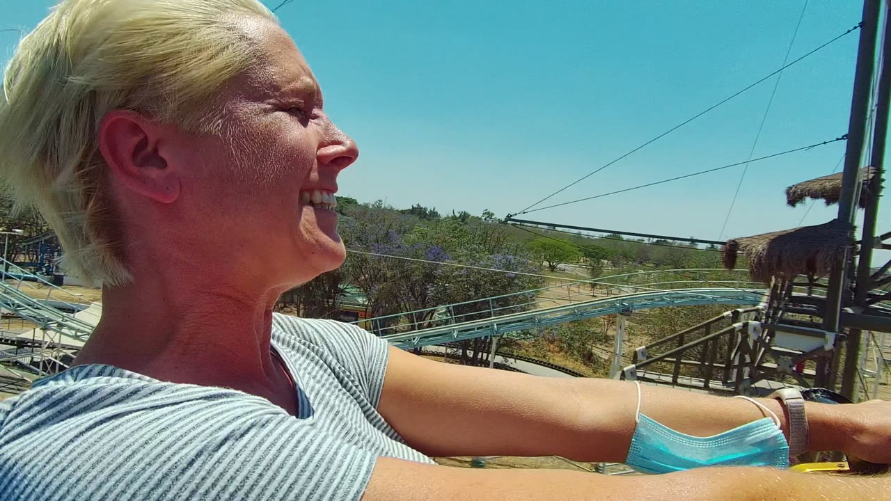 At 0:02, the woman grins while holding on during the ride, with roller coaster rails, utility lines, dry brush, and open blue sky visible around her.