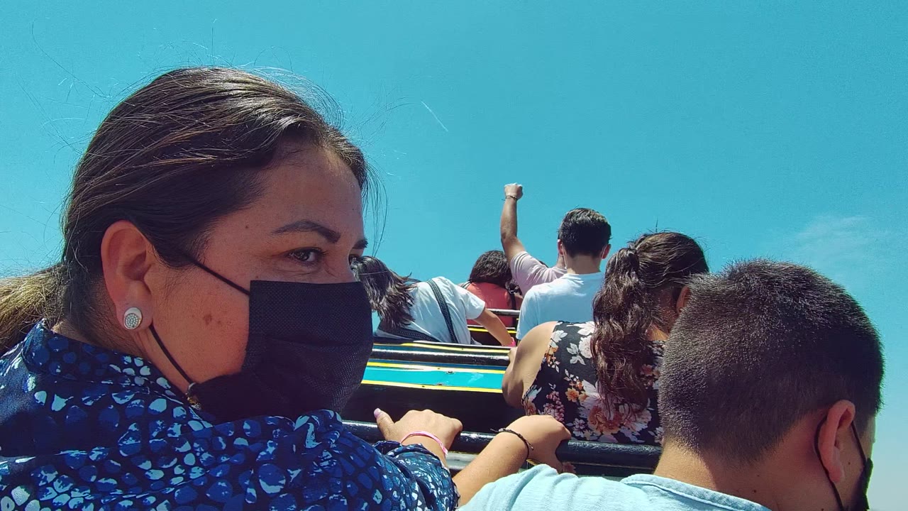 Foreground passengers, including a masked woman and a child, sit on a Guadalajara amusement ride with other riders ahead against a clear blue sky.