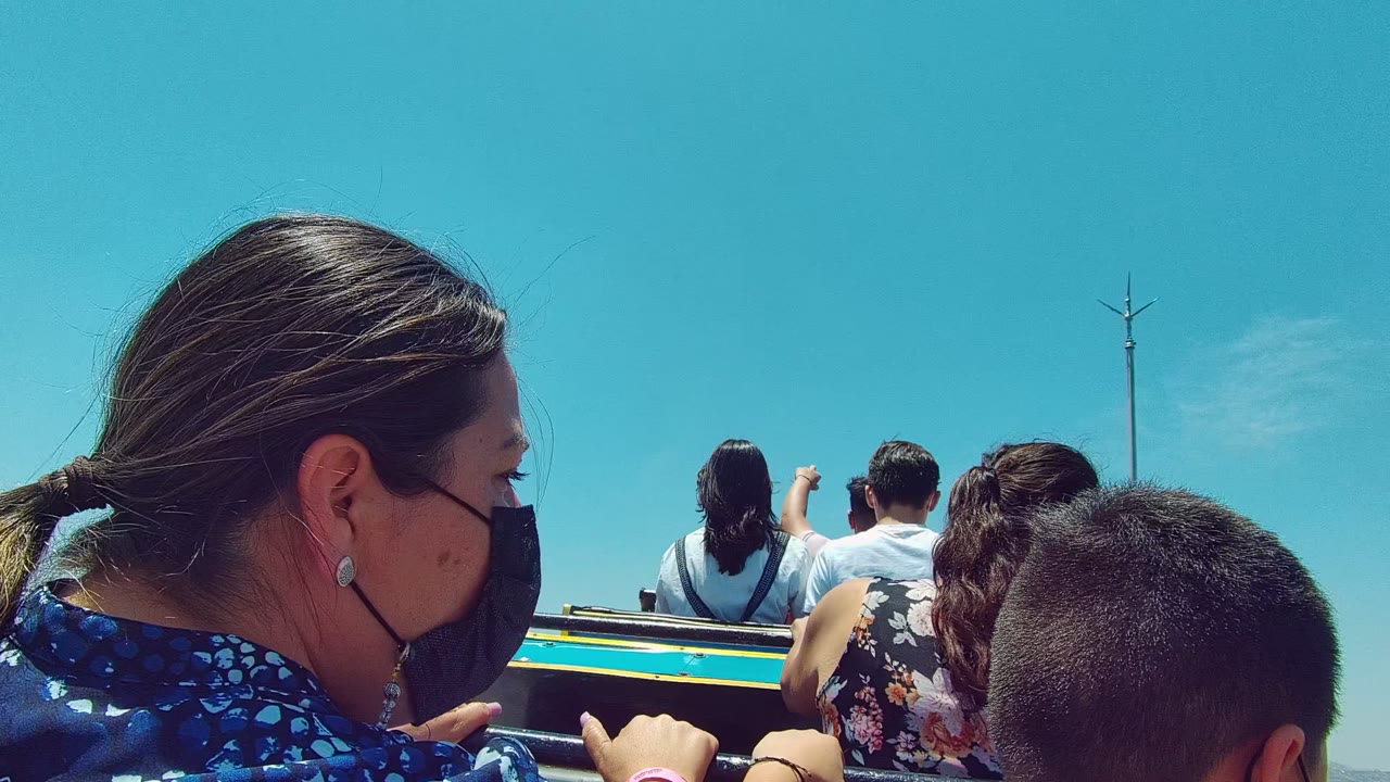 Foreground riders remain close to camera while passengers ahead raise hands and a tall pole appears against the blue sky in Guadalajara.
