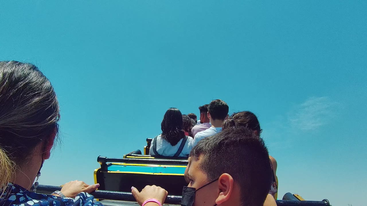 In Guadalajara Mexico 2022 footage, masked passengers sit in rows on an open vehicle, with a woman partly visible at left, a child in the foreground, and a clear blue sky above.