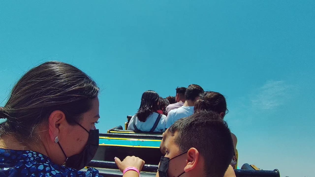 The masked woman turns into profile beside the child, with riders ahead, hands on the rail, yellow vehicle trim, and open blue sky in the Guadalajara Mexico 2022 clip.