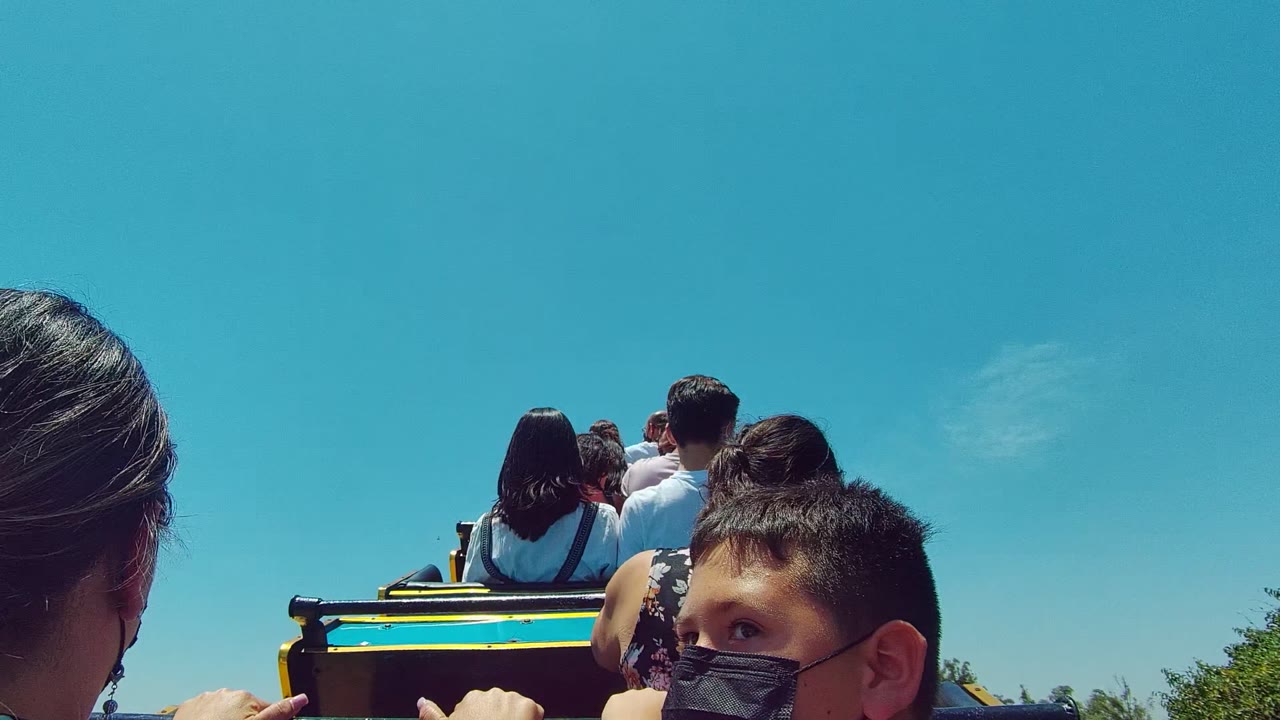 At 0:00, passengers on an amusement park ride in Guadalajara Mexico are framed from behind, with a masked boy partly visible at lower right and a clear blue sky filling the background.