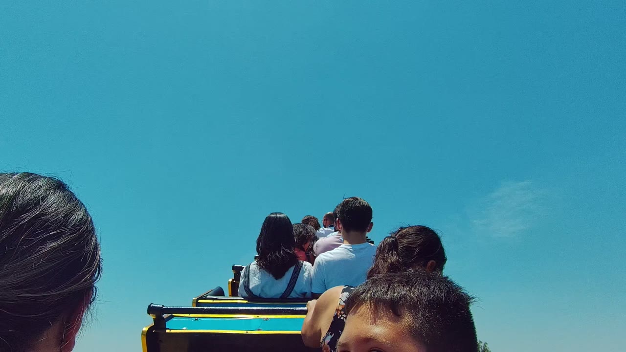 At 0:02, riders sit in rows ahead of the camera on the Guadalajara Mexico amusement ride, with the masked boy cropped at the lower right and the sky dominating the frame.