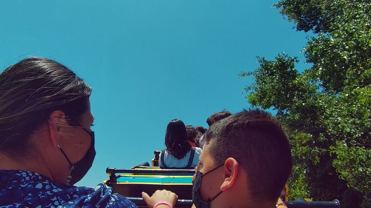 In Guadalajara Mexico, a masked woman and boy sit in the foreground of a colorful trolley ride with other passengers ahead and trees framing the blue sky.