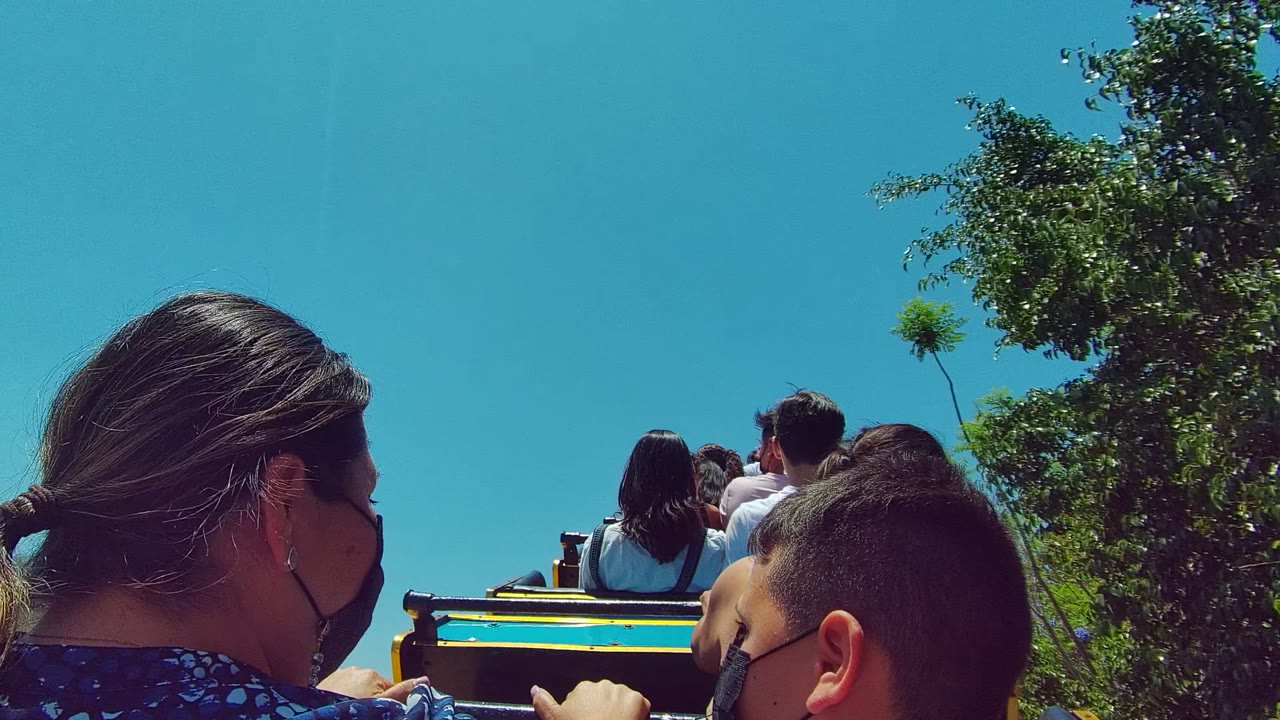 In Guadalajara Mexico, the camera looks past a masked woman and boy toward seated passengers on the trolley, with leafy trees on the right and clear blue sky overhead.
