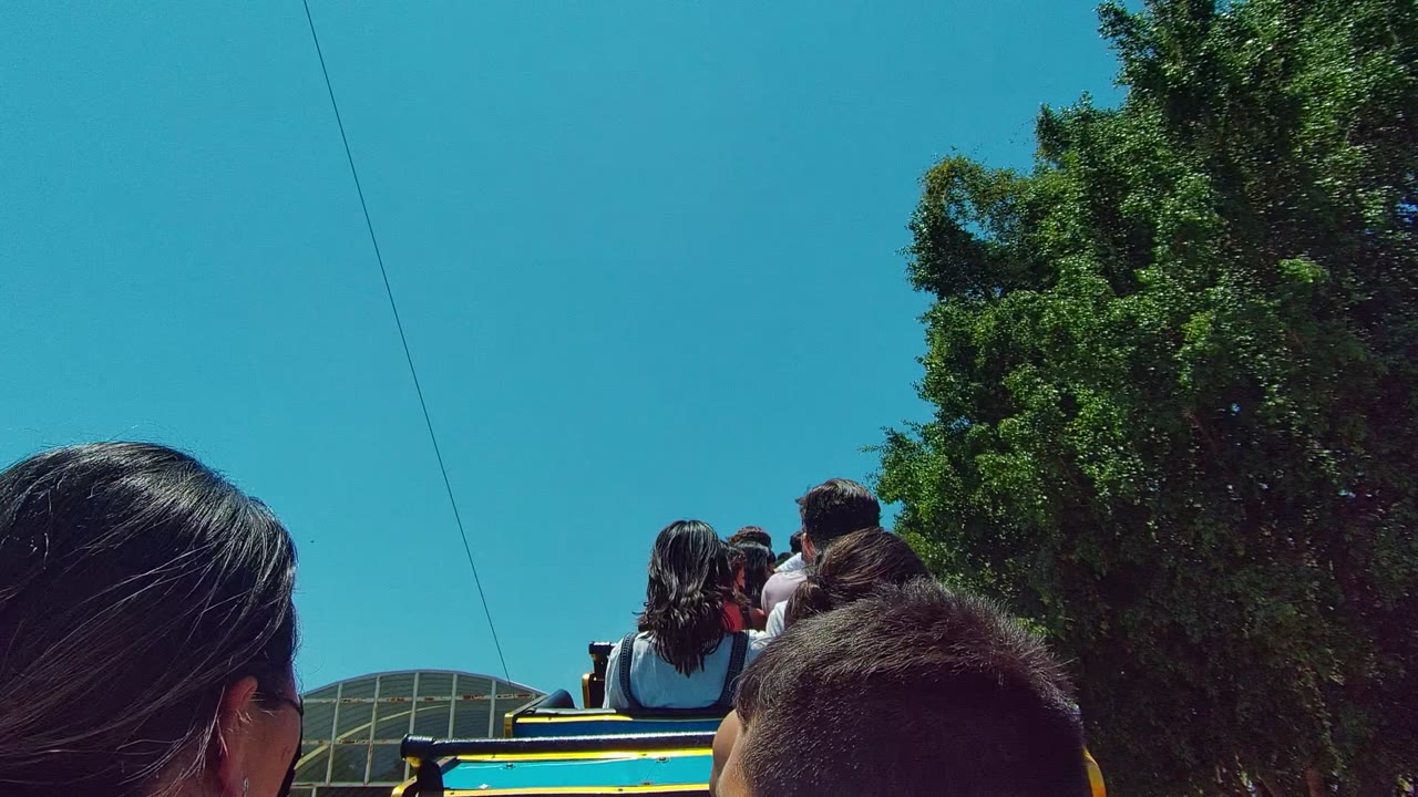 Passengers ride on an open-top vehicle in Guadalajara Mexico, with the back of a woman's head, a child's head, trees, blue sky, an overhead wire, and a curved structure visible.