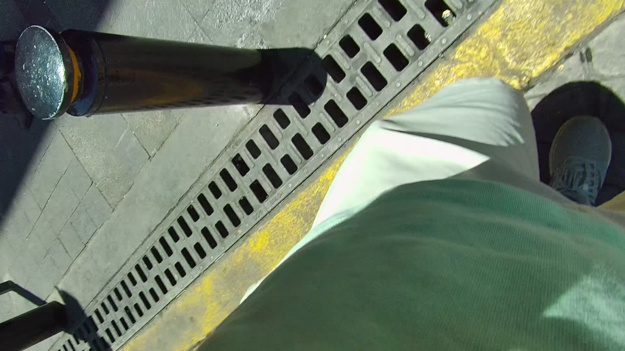 Downward low-angle frame in Mexico showing sidewalk paving, a metal drain grate, a yellow curb line, a black post, and a pedestrian's lower body near the camera.