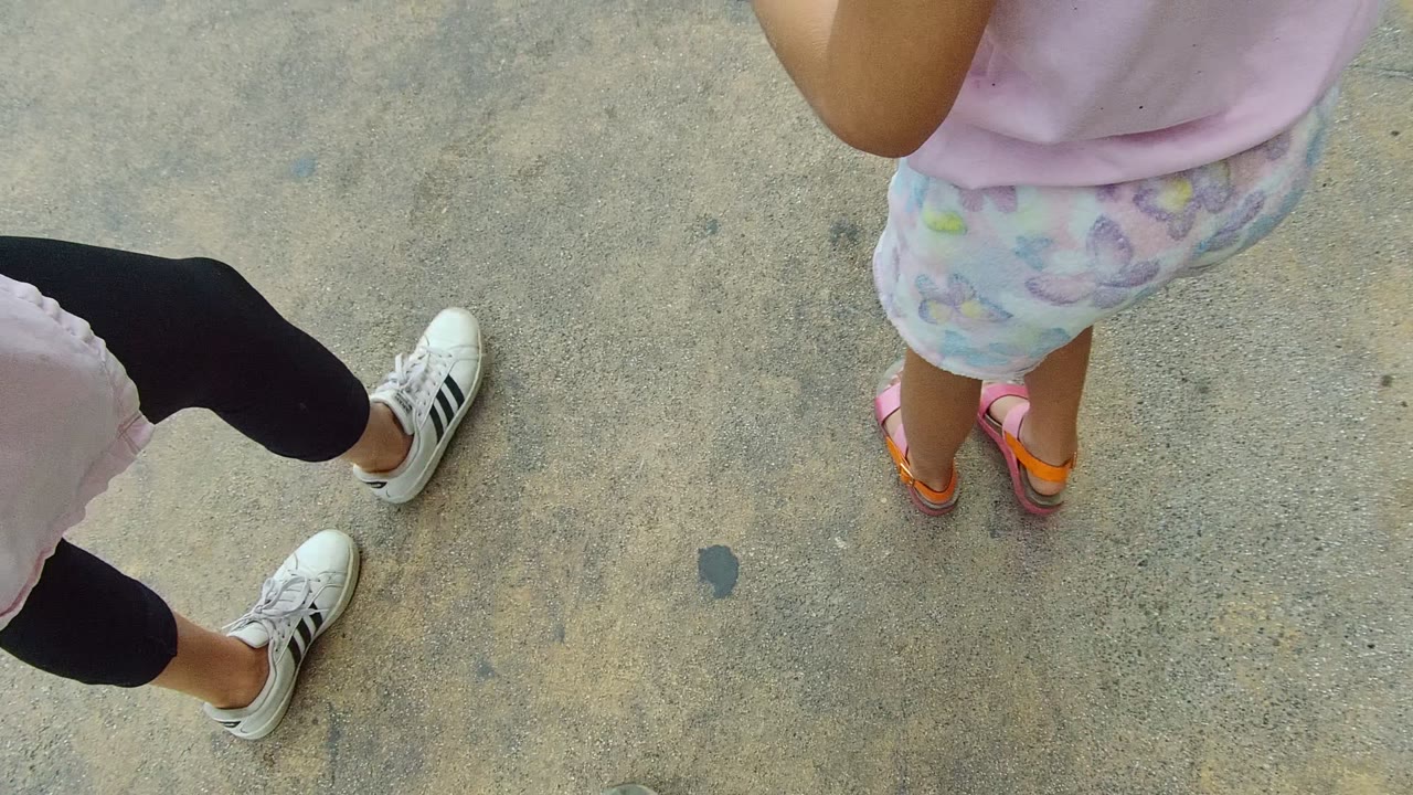 Low-angle Guadalajara Mexico frame showing pavement, an adult in a pale pink dress and white sneaker, and a child stepping in pink sandals.