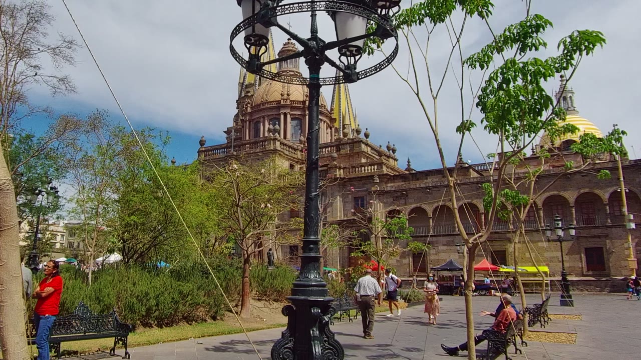 A Guadalajara, Mexico public square frames a cathedral with domes and arches behind trees, benches, a black lamppost, and several visible people.