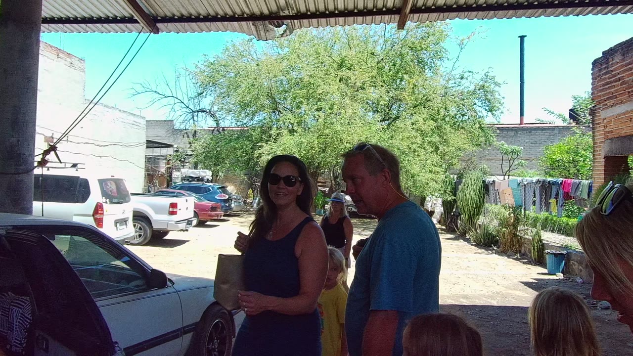 Adults stand under a corrugated roof beside a white car in a residential courtyard in Mexico, with parked vehicles, trees, laundry, and brick walls behind them.