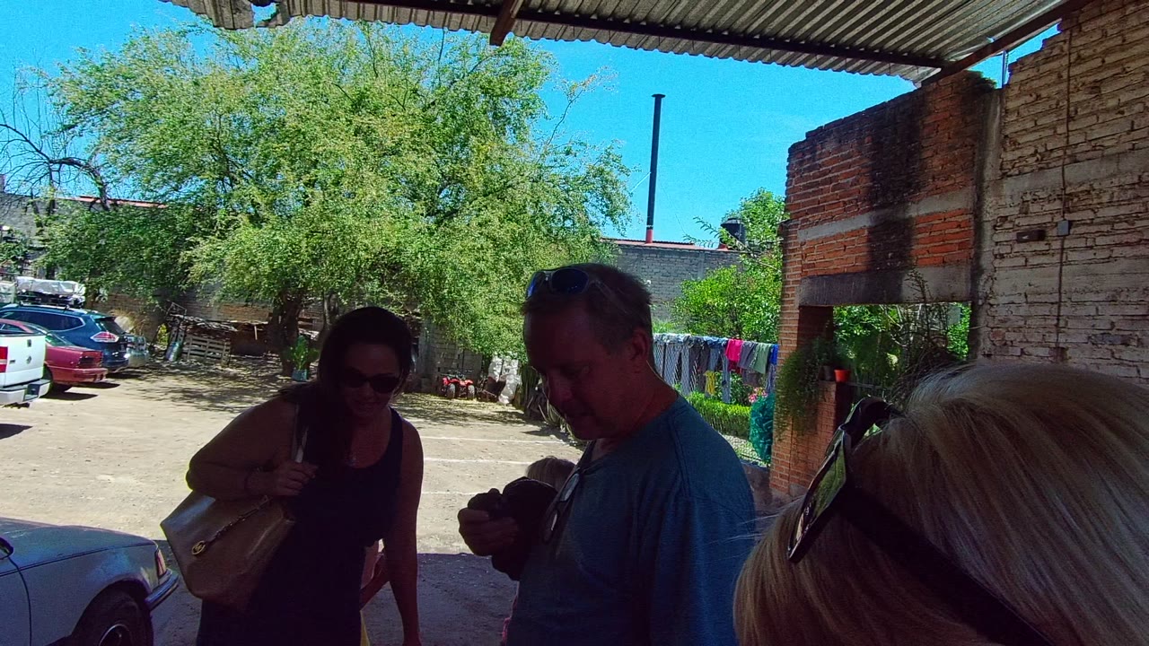 The woman adjusts or holds her shoulder bag in the shaded foreground while the man looks down, with a white pickup, trees, laundry, and brick masonry in the Mexico courtyard background.