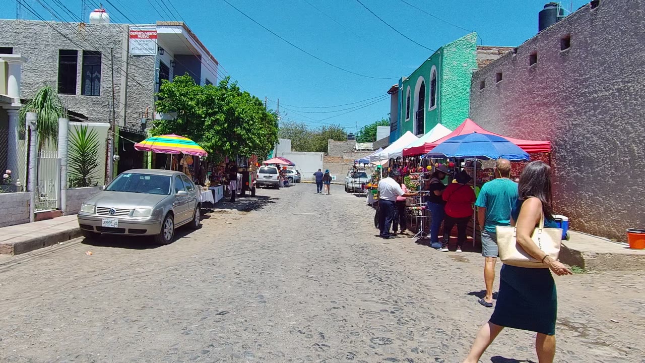 A sunny cobblestone street in Mexico shows market stalls under colorful umbrellas, parked cars, low buildings, and several pedestrians near the vendor tables.