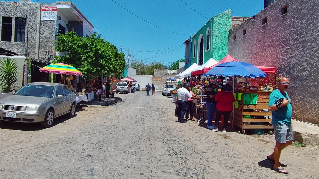 A man crosses the foreground of a sunny market lane in Mexico as shoppers stand by stalls under red, blue, and striped umbrellas beside parked cars.