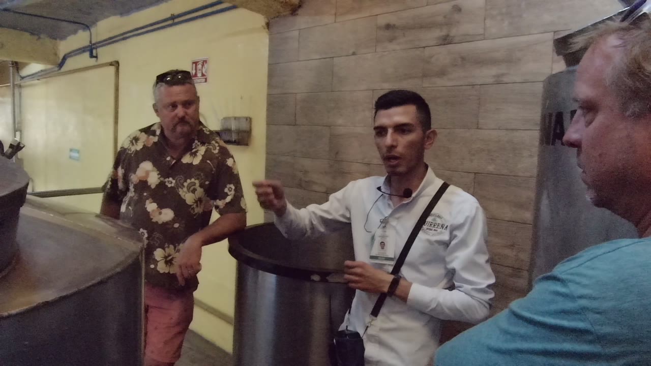 In Tequila, Jalisco, Mexico, three men stand beside large metal tanks as the man in the white shirt begins explaining something to two listeners.