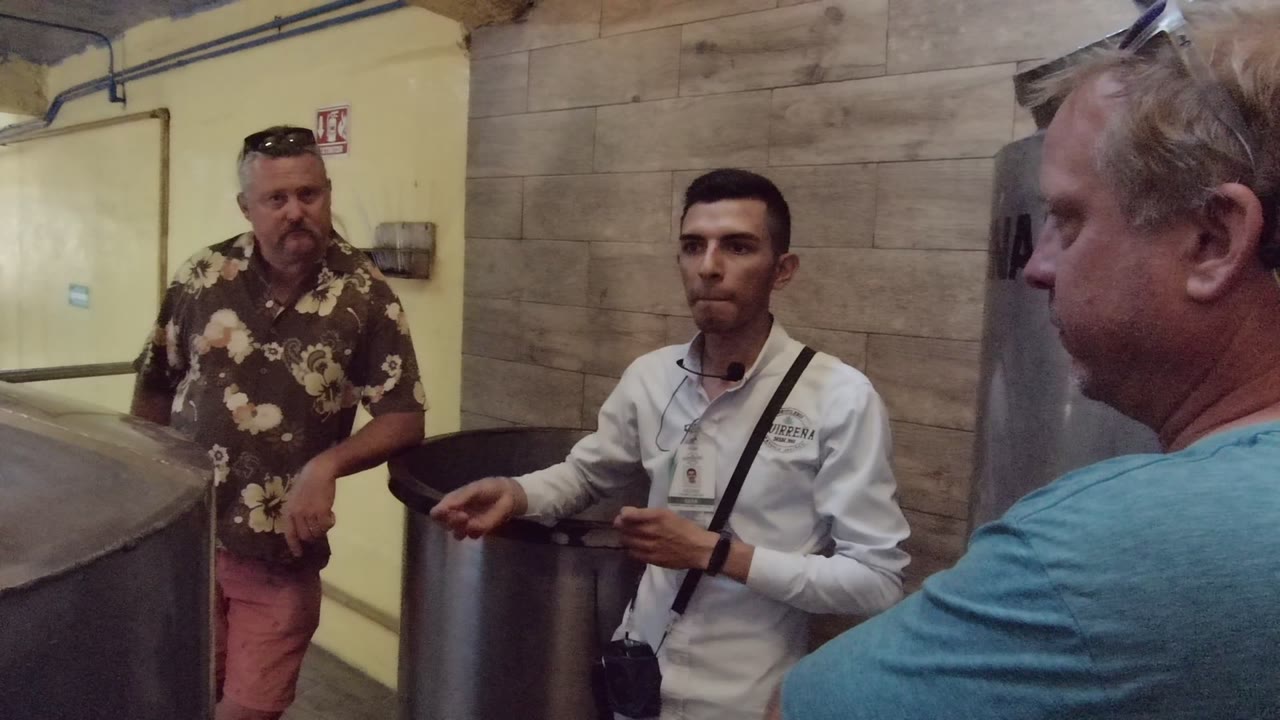In Tequila, Jalisco, Mexico, the speaker stands by the metal vessel with two men listening, including a close side view of the man in the blue shirt.