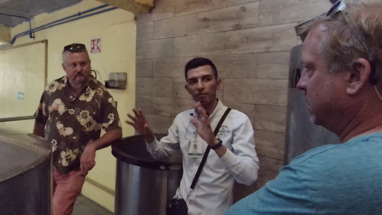 In Tequila Jalisco Mexico, a guide in a white shirt gestures while two adult male visitors stand near large metal tanks in an indoor industrial room.