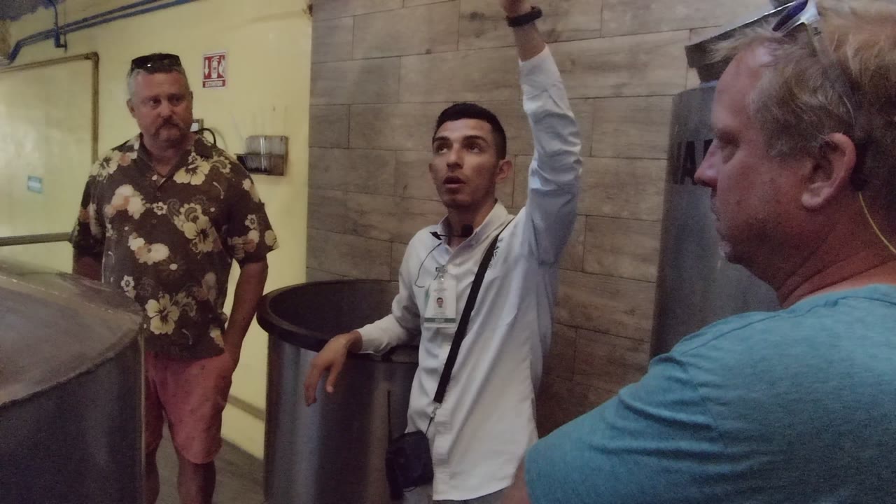 Three men stand beside stainless metal tanks inside an industrial room in Mexico, with the man in a white shirt raising one arm while the others watch.