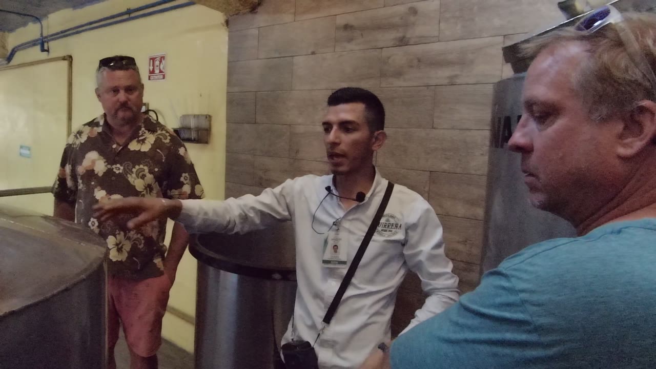 The white-shirted guide gestures across the tank area as the floral-shirted man faces him and the blue-shirted man appears in close foreground profile in Mexico.