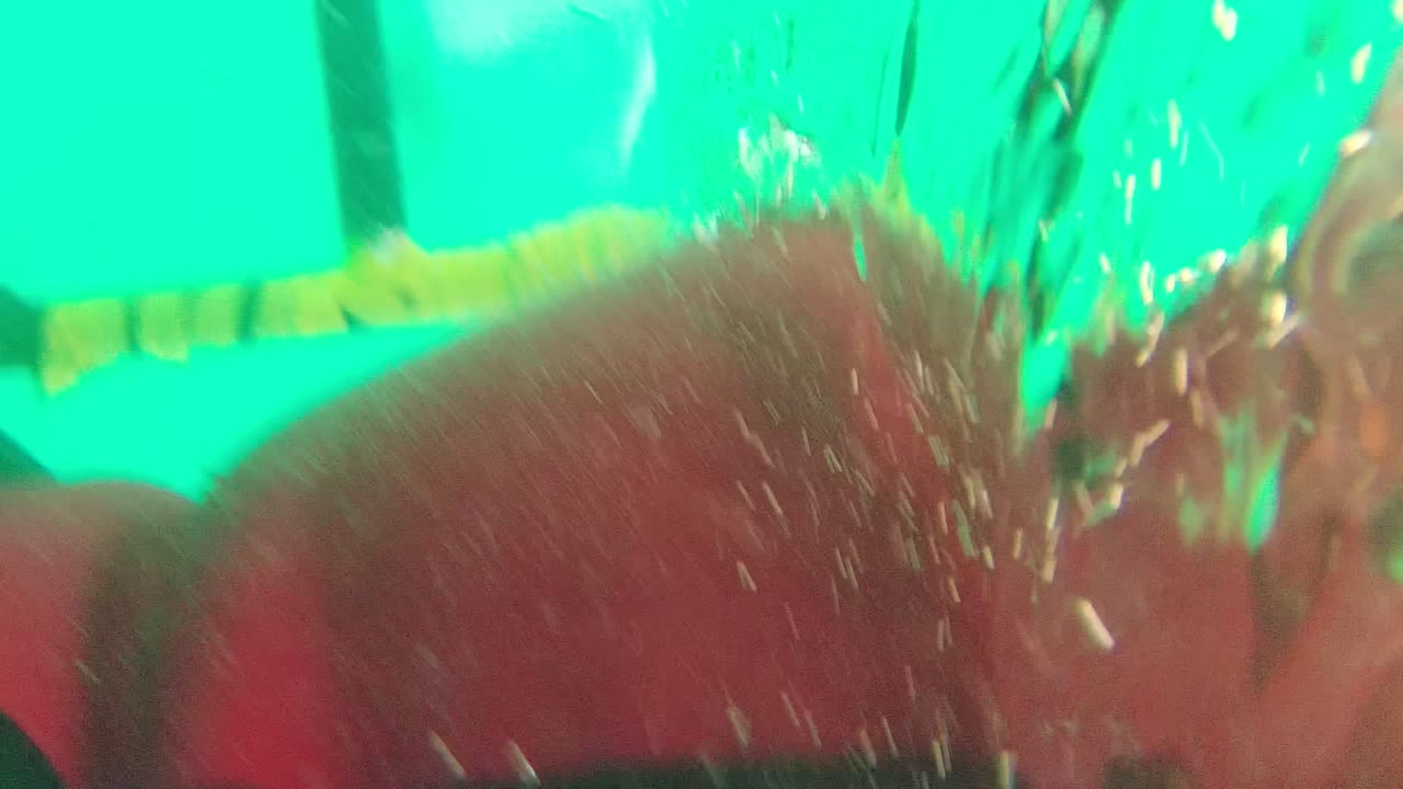 A swimmer in a red cap or red swim gear passes very close to the underwater camera, with bubbles and a yellow and black lane divider behind them in Mexico.