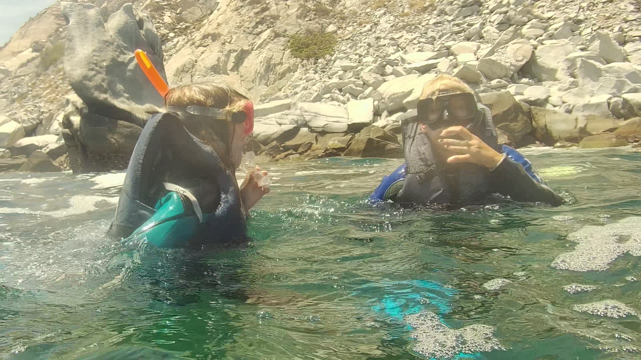 Two snorkelers at the surface with fins and rocks behind them.
