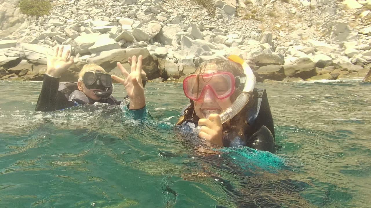 Two snorkelers float at water level in Cabo Pulmo, Mexico, with one person raising both hands near the camera and a pale rocky shoreline behind them.