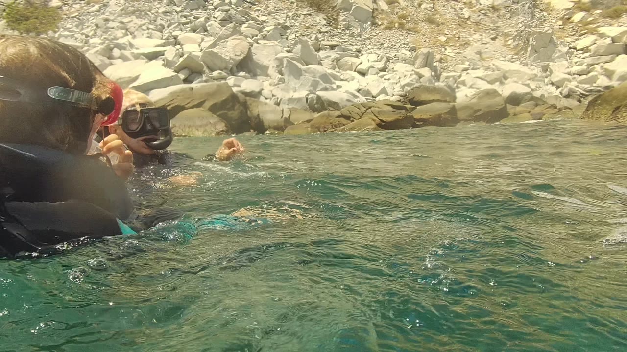 Two snorkelers in Cabo Pulmo, Mexico, are seen from water level as one turns sideways in the foreground and the other stays farther back near the rocky shore.