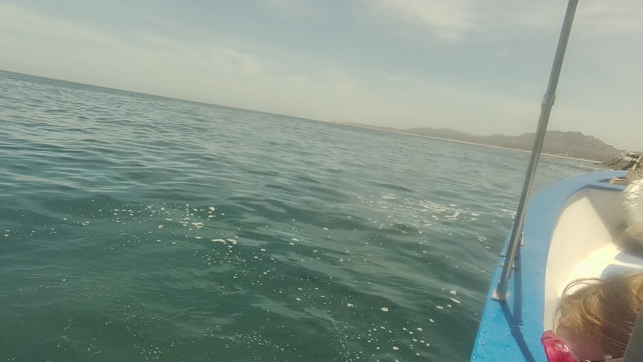 Canted opening frame from Cabo Pulmo, Mexico, showing green-blue ocean, distant shoreline, a vertical boat pole, and the blue edge of a small boat with part of a person just visible at the lower right.