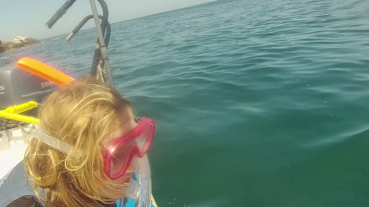 A child wearing a pink snorkel mask stands near the edge of a boat in Cabo Pulmo, Mexico, with green-blue ocean water and boat equipment behind them.
