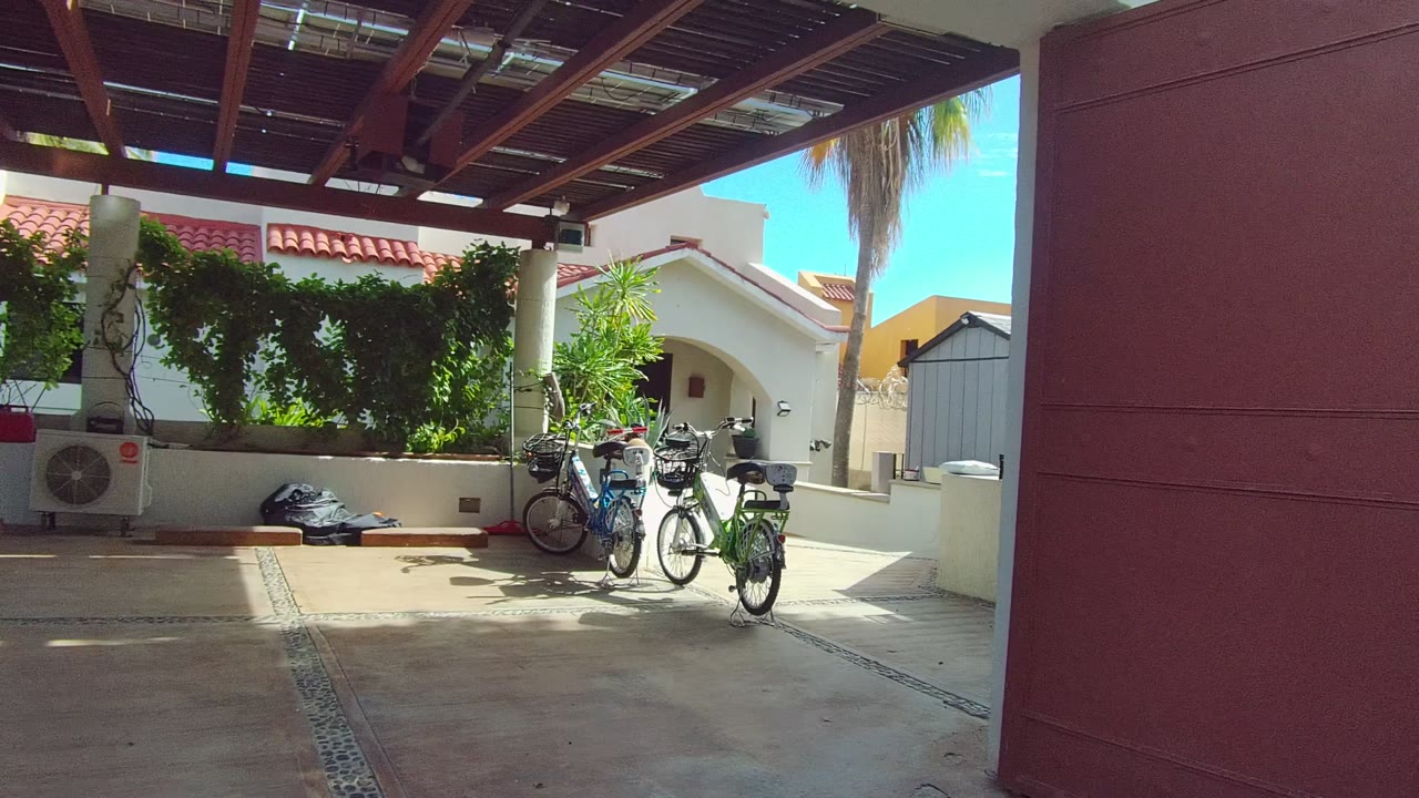 Opening frame of a covered carport in East Cape Los Cabos BCS Mexico with bicycles parked near white columns, green vines, a red door at right, and no visible people.