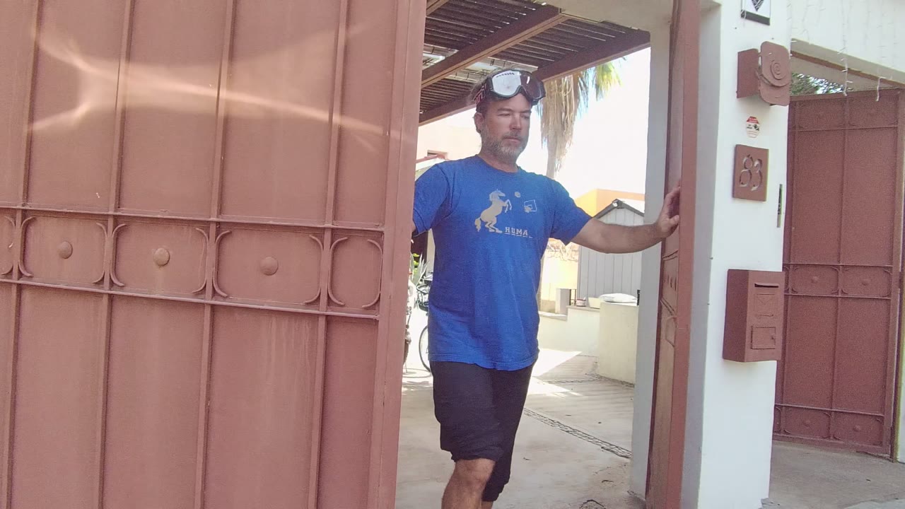 A man in a blue shirt stands partly behind an open red metal gate at a residential courtyard in East Cape Los Cabos, Mexico.