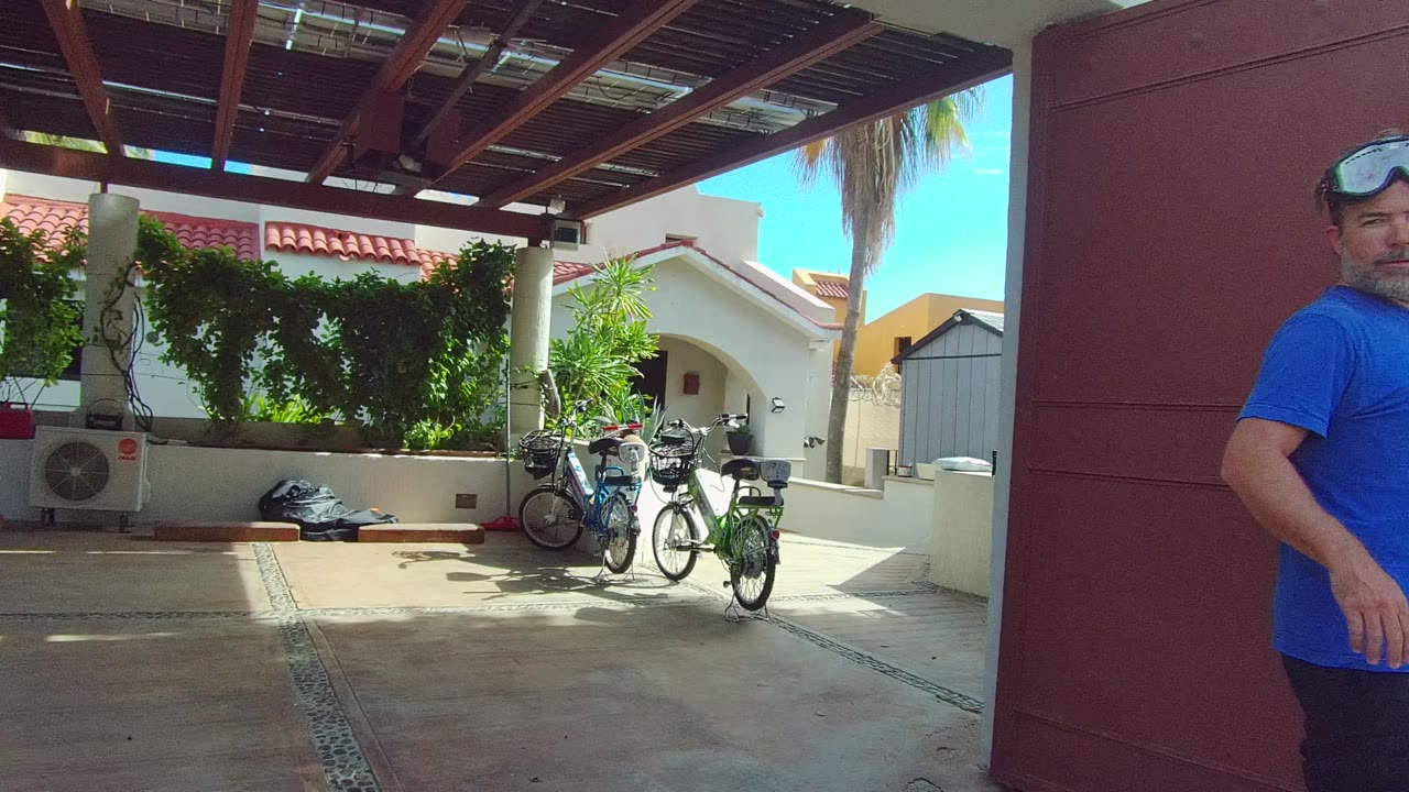 A man in a blue shirt stands beside the red metal gate with bicycles, a shaded roof structure, white columns, and greenery behind him in East Cape Los Cabos, Mexico.