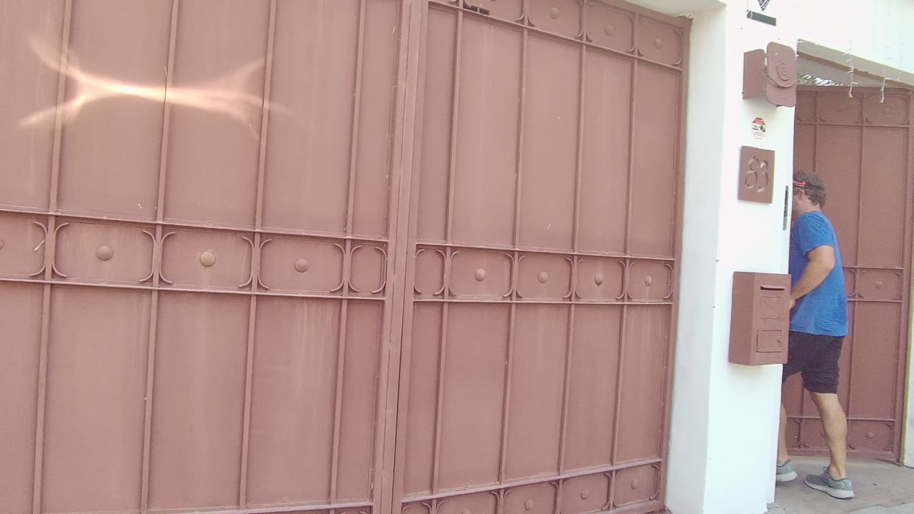 Closed reddish brown metal gate at a residential entrance in East Cape Los Cabos, Mexico, with white walls, a wall-mounted box, bright daylight, and no visible people.