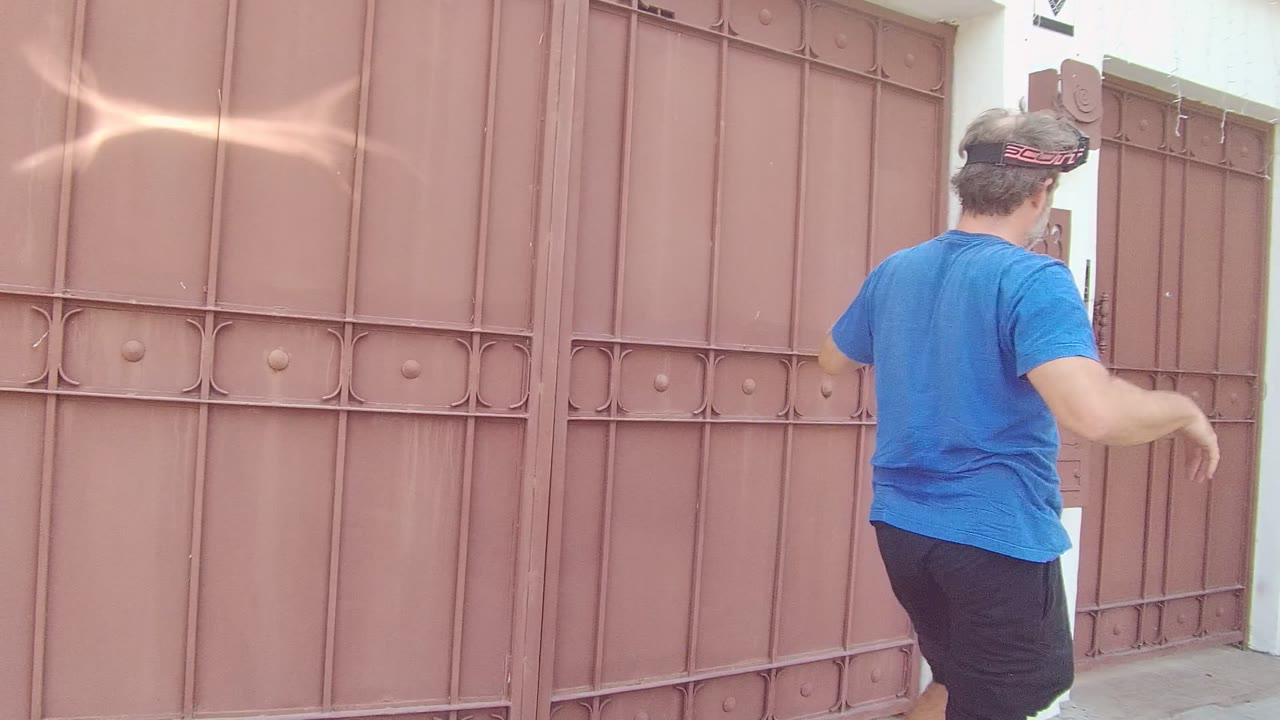 In East Cape Los Cabos, Mexico, a man in a blue shirt stands near the right side of a large brown metal gate beside a white wall and access panel.