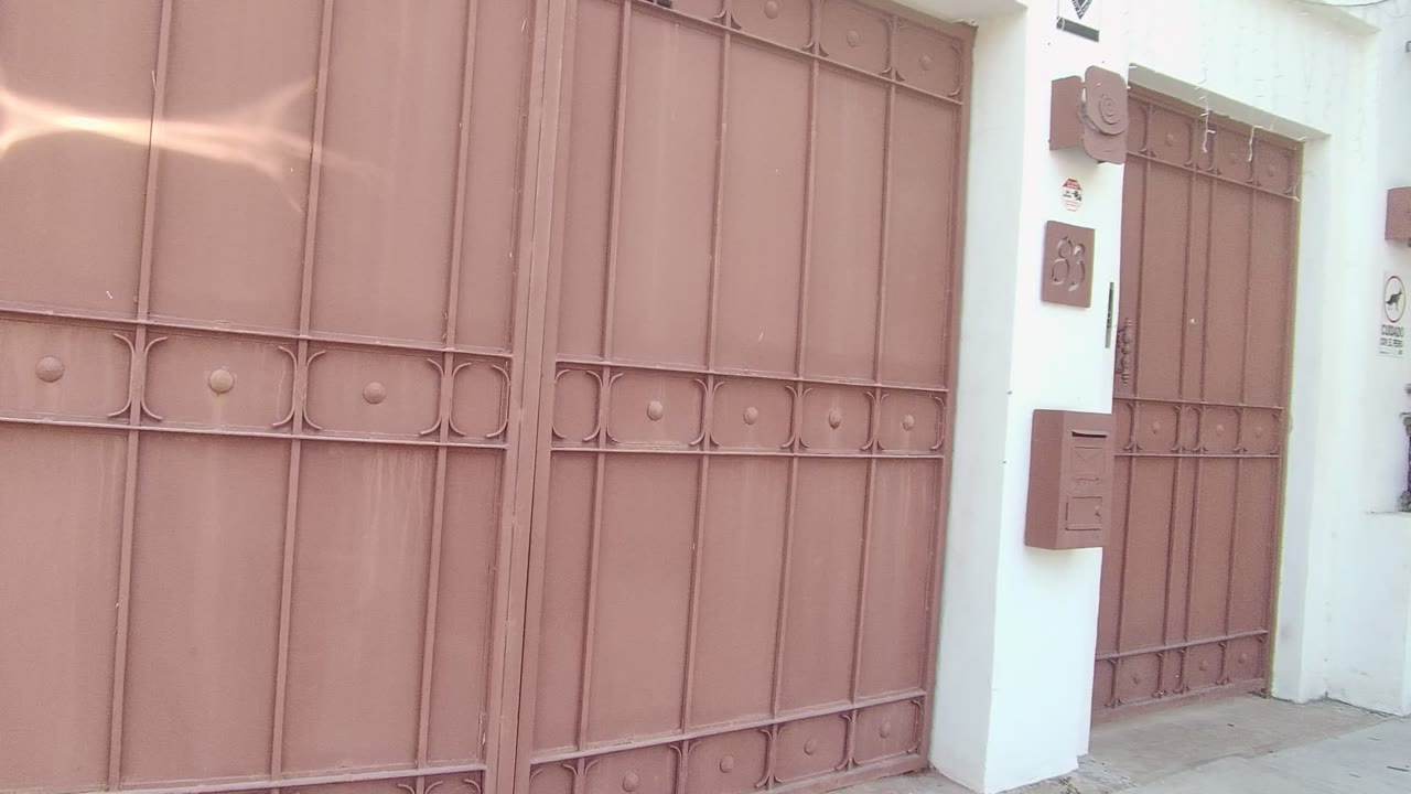 Closed brown metal gate in East Cape Los Cabos, Mexico, with a white wall, side doorway, mailbox, intercom, and no visible people at the start of the clip.