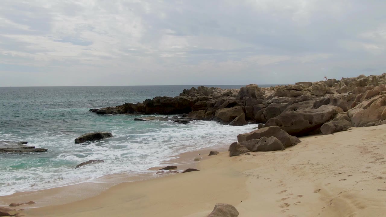 Wide view of Shipwrecks East Cape BCS Mexico beach with pale sand, rounded rocks, surf breaking at the shoreline, cloudy sky, and no visible people.