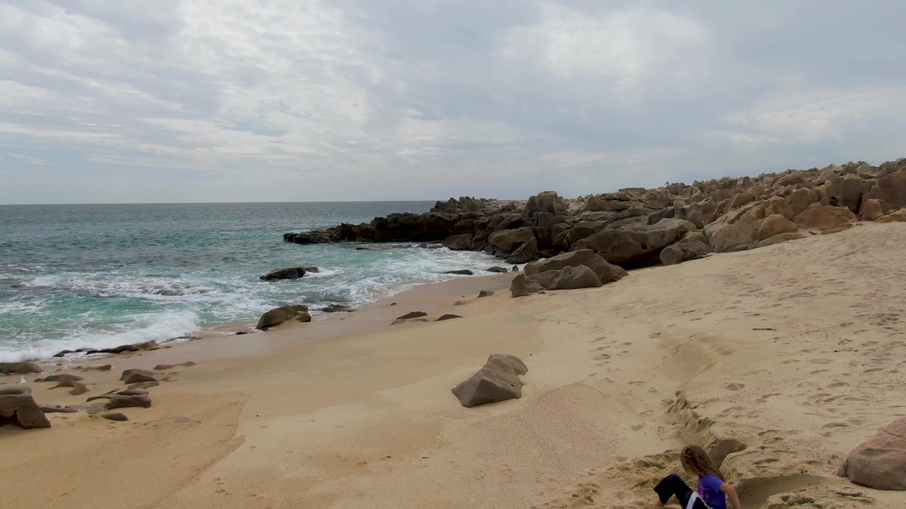 Opening frame of Shipwrecks East Cape BCS Mexico beach with tan sand, blue-green waves, dark rocks, layered clouds, and a child in a blue shirt partly visible along the lower edge.