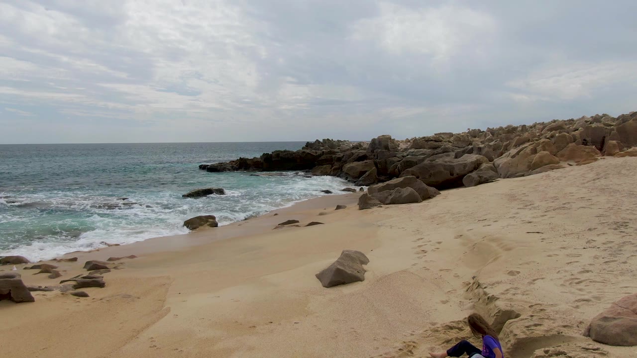 Ocean waves break beside rocks while a child remains partly visible near the lower beach edge.
