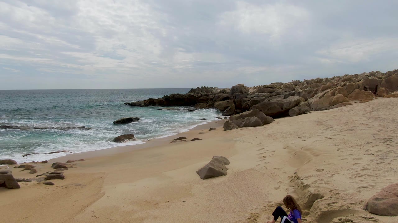 Third frame holds on the Shipwrecks East Cape BCS Mexico shoreline, with foamy surf at left, rounded rocks at right, and the child cropped near the lower right corner.
