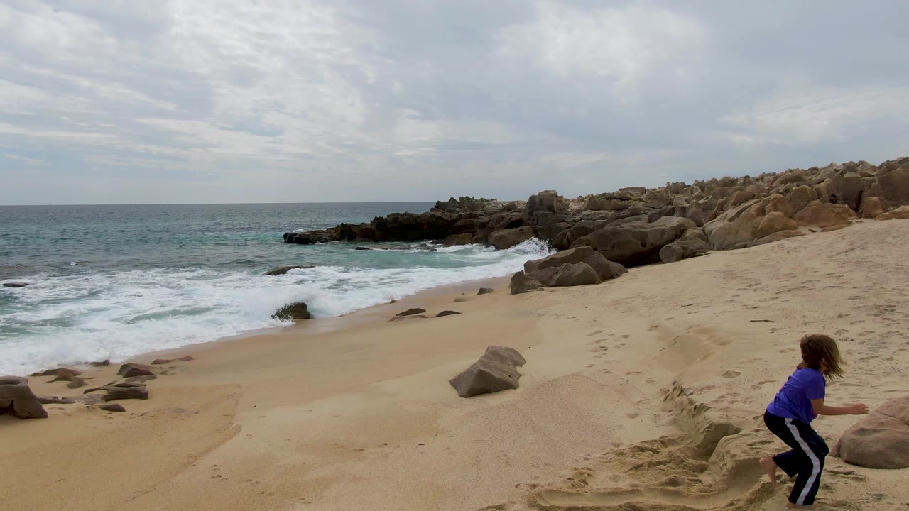 At Shipwrecks East Cape BCS Mexico, a child in a purple shirt crouches near a sandy edge while waves break beside dark rocks under a cloudy sky.