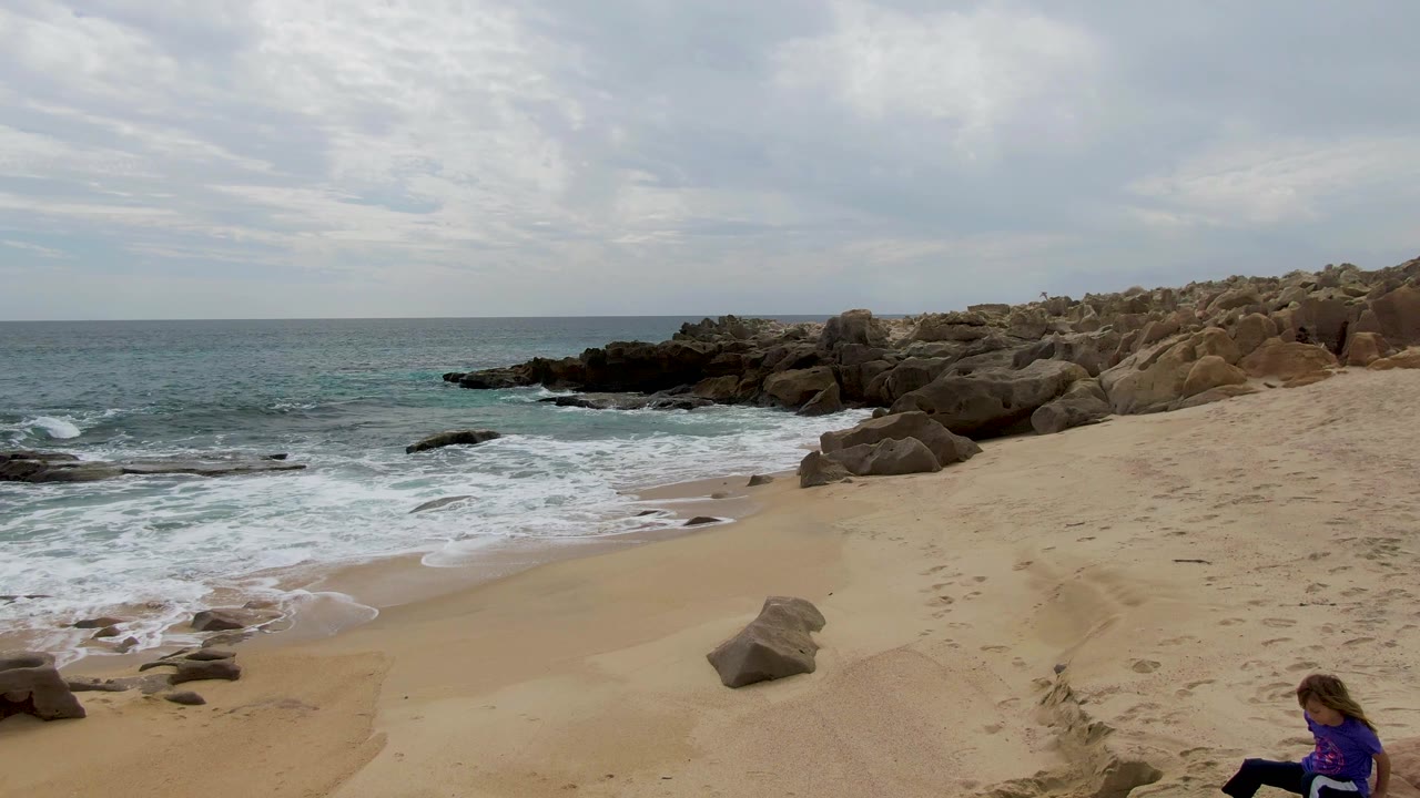 Opening frame at Shipwrecks East Cape BCS Mexico showing pale sand, foamy surf, tan boulders, an overcast sky, and a person in a purple shirt partly visible at the lower right.