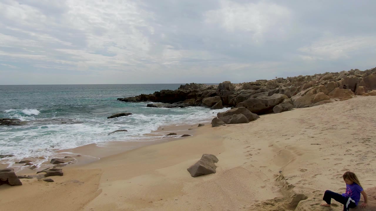 Foamy ocean waves wash toward a sandy beach beside rocks as a seated person appears near the lower right.